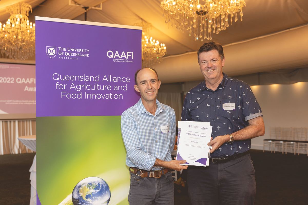 Two men smiling as one presents the other with an award certificate under chandeliers