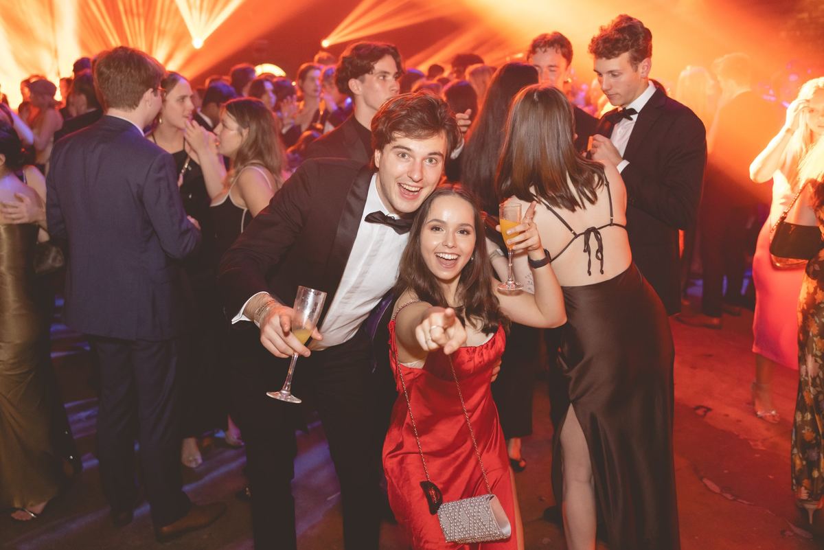 A guest in a tuxedo and a friend in a red dress pointing at the camera on a packed ball dance floor