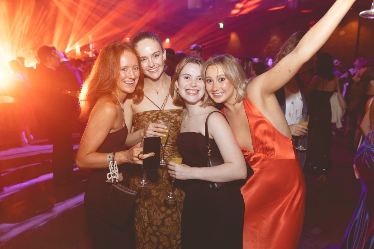 Four friends in evening gowns smiling together on a crowded dance floor under orange stage lights