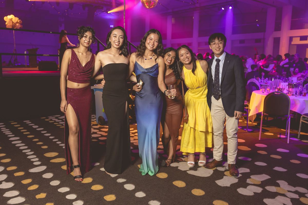 Six friends in formal wear posing together beside the dance floor, dinner tables and purple lights behind them