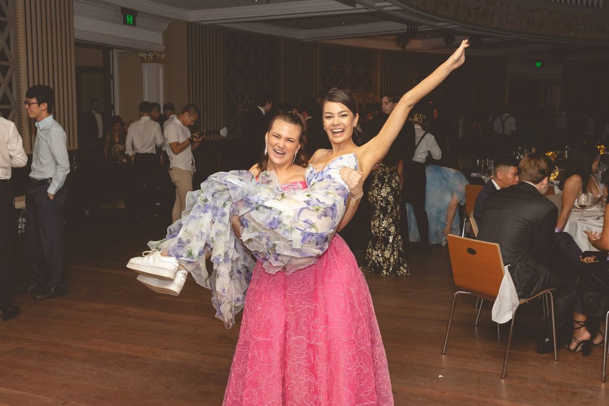 A guest in a pink gown lifting her laughing friend off her feet in a busy ballroom