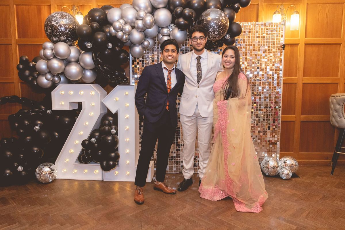 Three guests in front of light-up 21 letters and a black and silver balloon arch