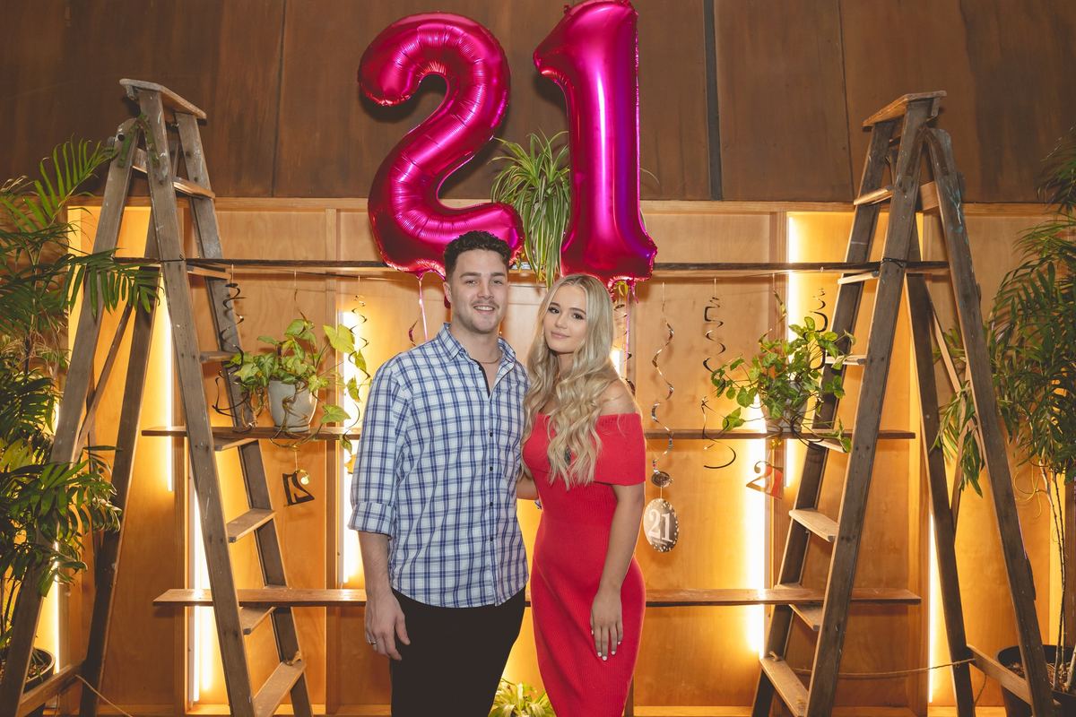 Two guests standing under giant pink 21 balloons between two wooden ladders strung with lights