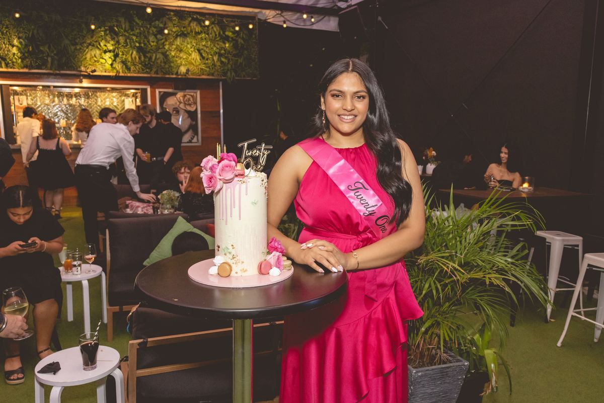 A woman in a pink dress and a Twenty One sash standing beside her birthday cake