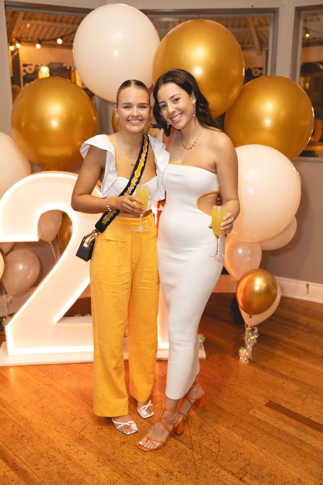 Two friends with drinks in front of gold and cream balloons and a light-up number