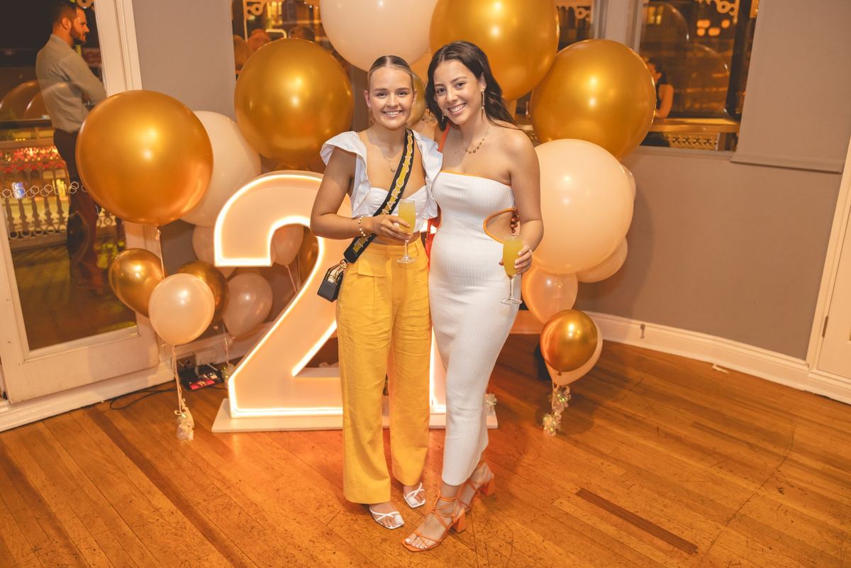 Two friends with drinks in front of gold and cream balloons and a light-up number