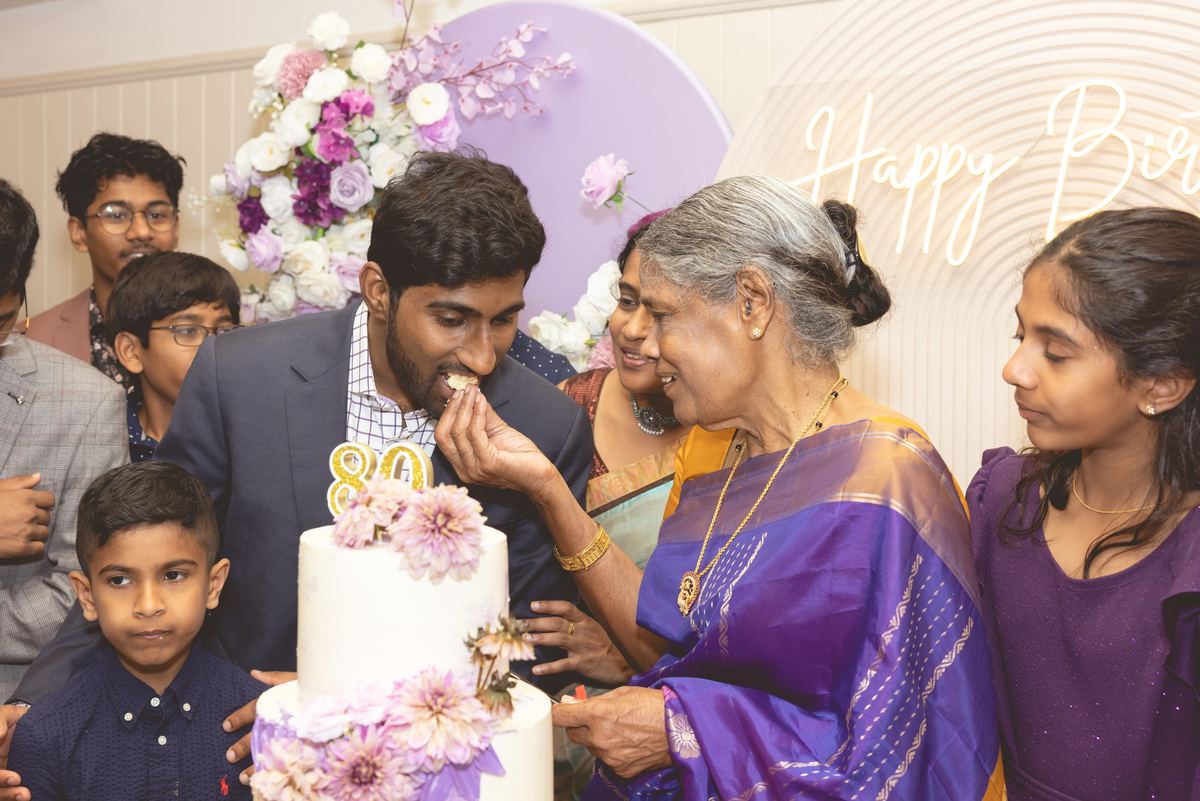 An older woman in a purple sari feeding cake to a young man beside an 80th birthday cake, family around them