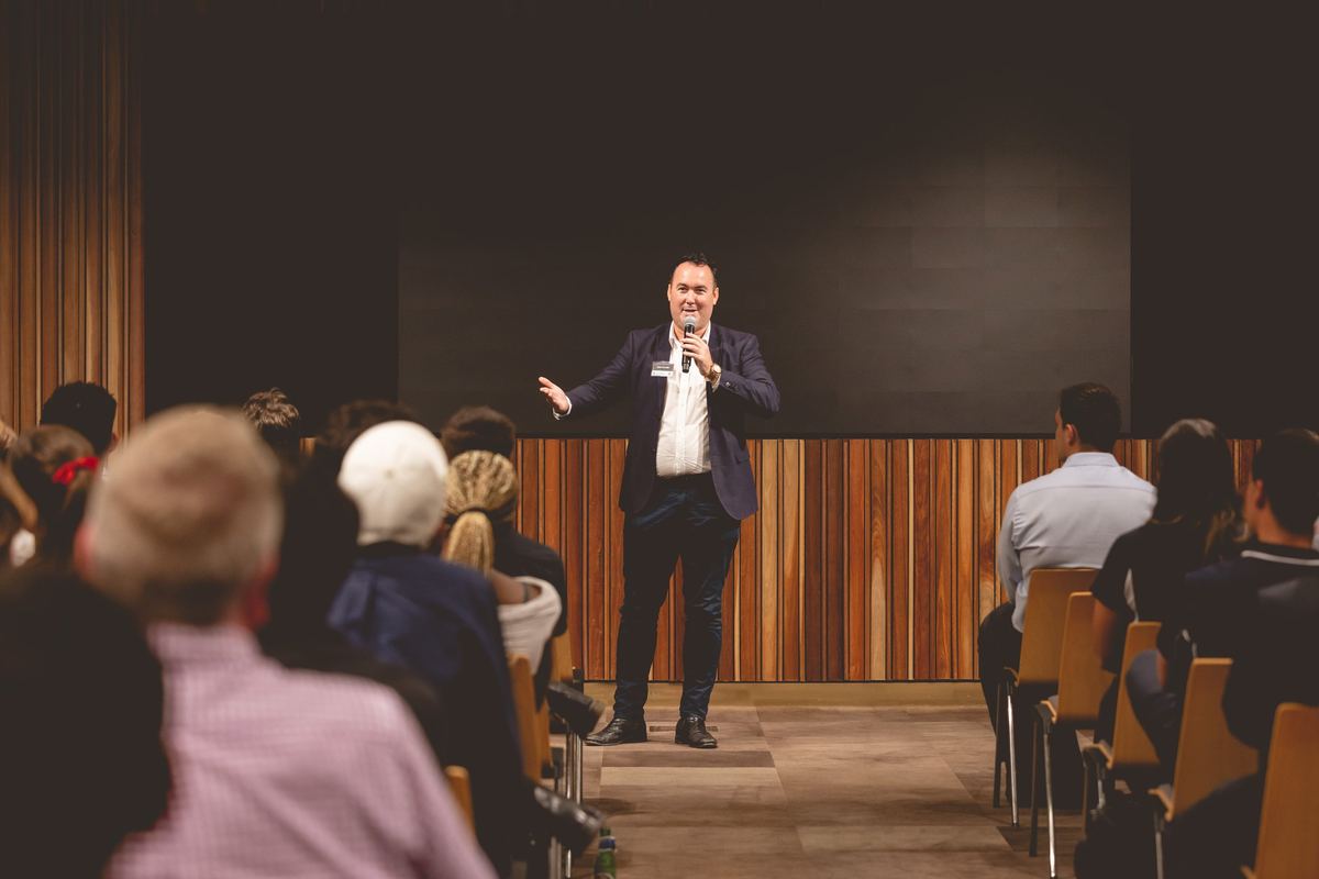 A speaker with a microphone talking to a seated audience from a timber lined stage