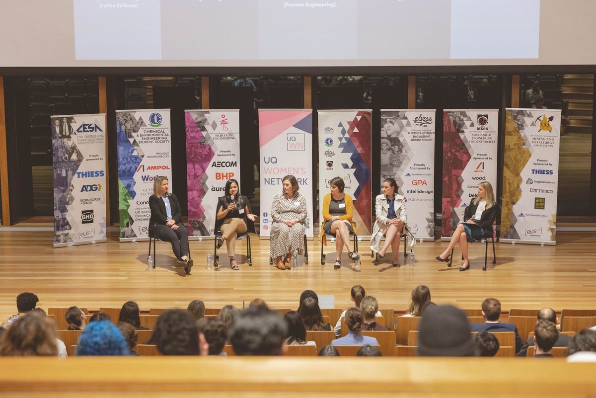 Six panellists seated on a stage in front of sponsor banners, the audience in the foreground