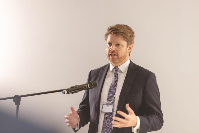 A speaker in a suit talking at a microphone and gesturing with both hands
