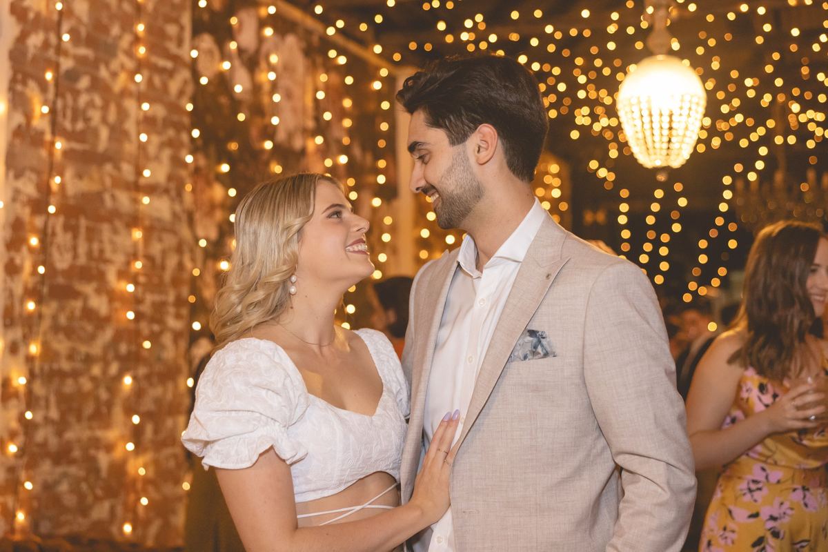 A couple smiling at each other under strings of fairy lights at their celebration