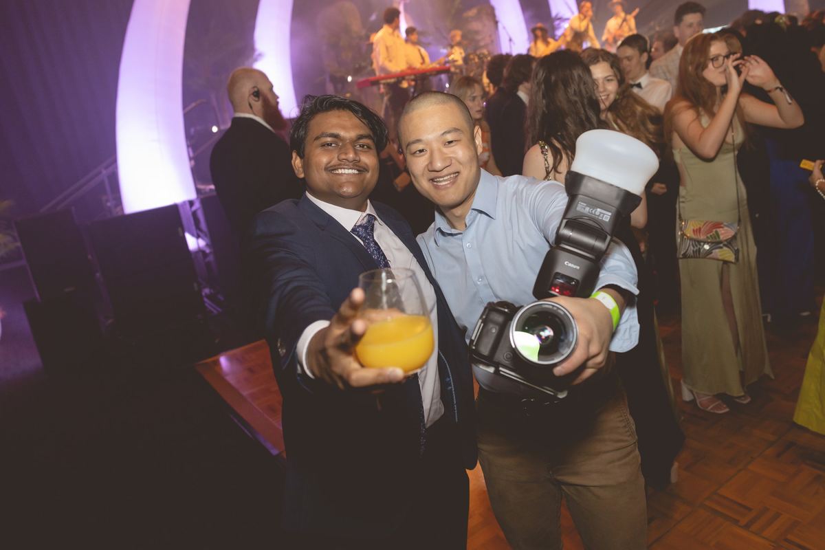 A photographer with a camera and flash posing with a guest who is raising a drink at a ball