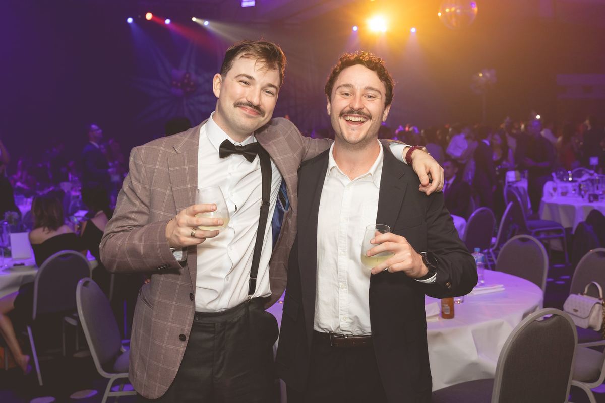 Two friends in suits, one in a bow tie, grinning with drinks among the dinner tables