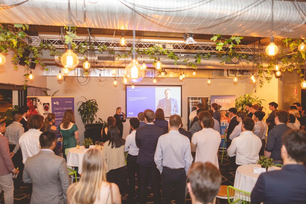 Guests in business wear under hanging bulbs and vines, watching a presentation on the screen