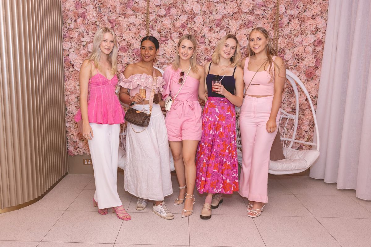 Five friends in pink and white outfits posing in front of a wall of pink roses