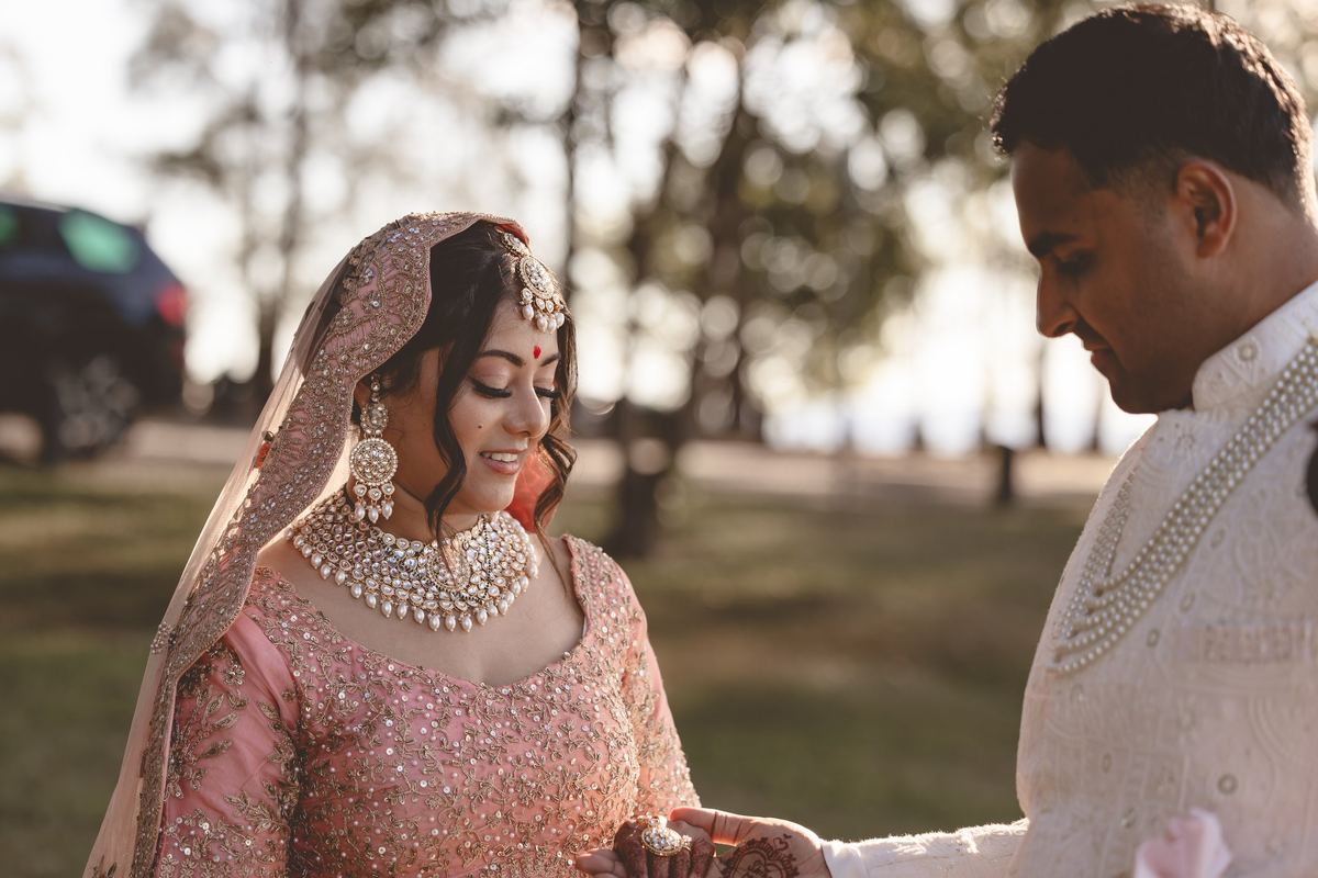 A bride in gold and pearl jewellery and a pink veil smiling down at her hands beside her groom
