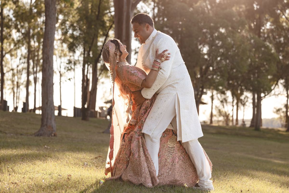 A groom in white dipping his bride in her embroidered pink gown under trees in golden evening light
