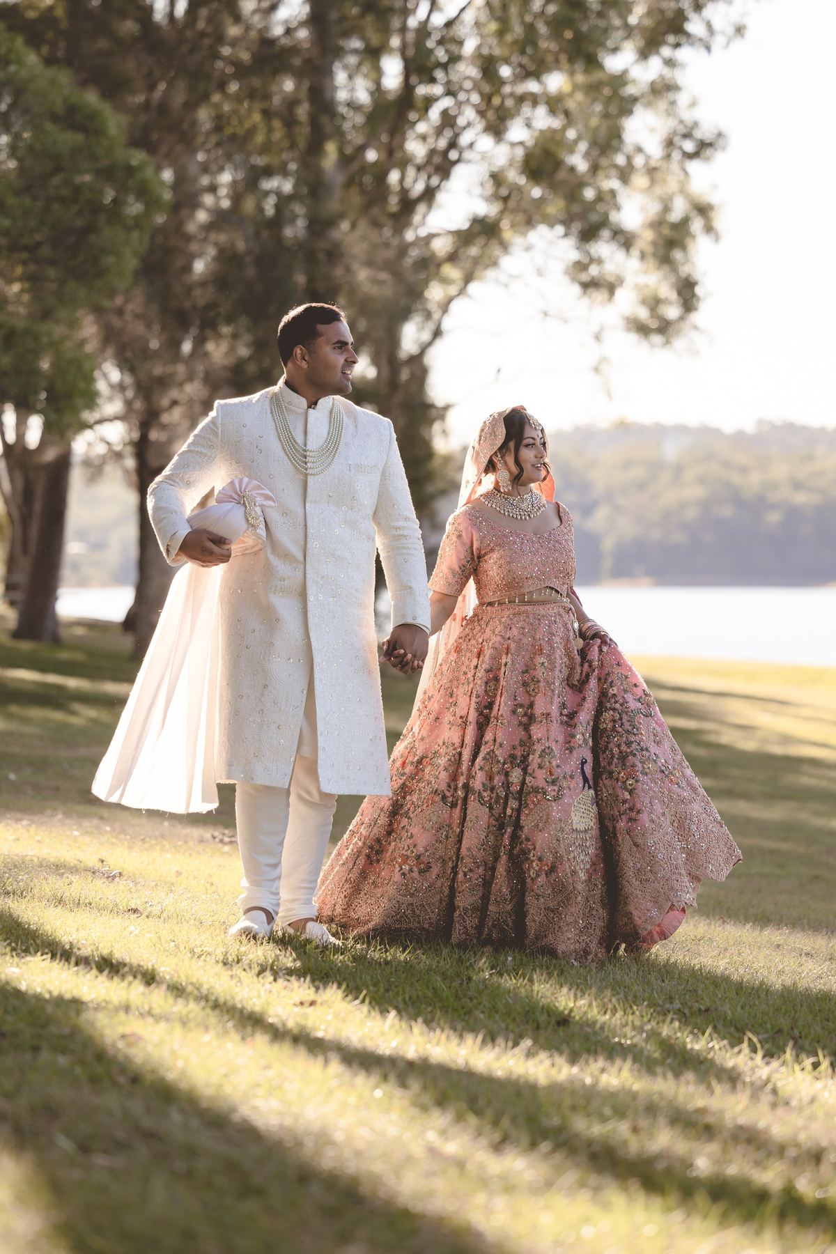 A bride and groom walking hand in hand across the grass by the water in low sun