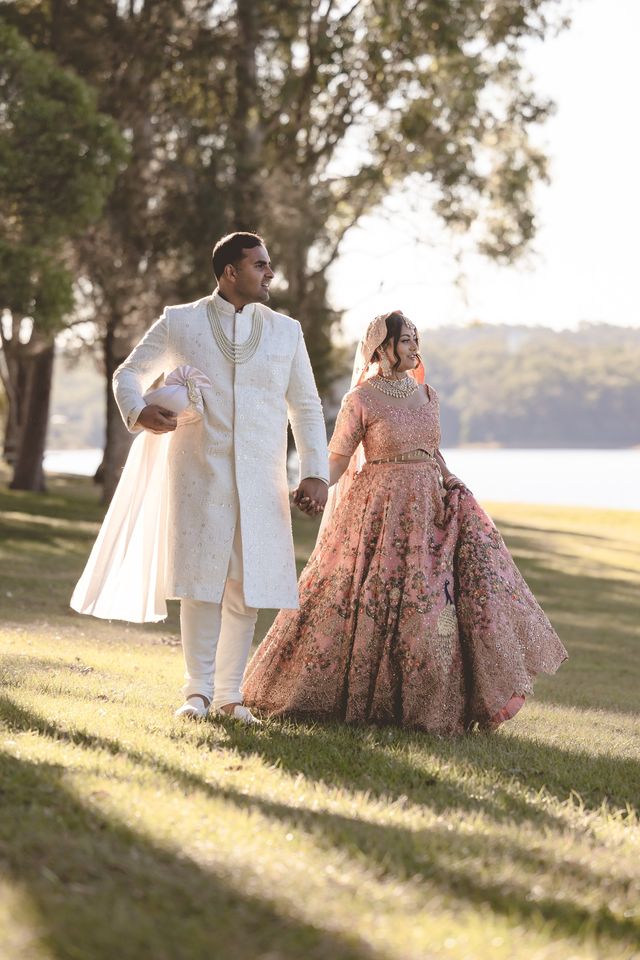 A bride and groom walking hand in hand across the grass by the water in low sun